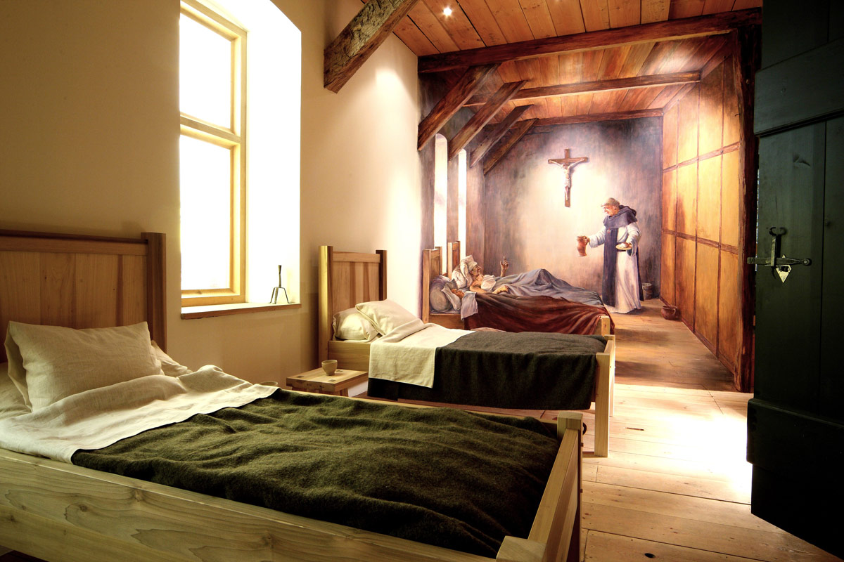 Twee bedden in de Ziekenzaal van het Klooster. Op de achtergrond is een schildering te zien van een kruisheer die iemand verzorgt.